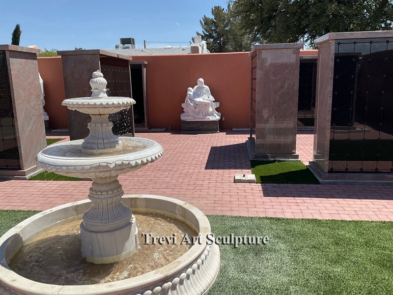 Tiered pale stone fountain on church grounds with religious sculpture behind